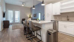 Una cocina moderna con armarios blancos, encimera de granito y electrodomésticos de acero inoxidable. Se observa un comedor con mesa y sillas, además de un cubo de basura pequeño.