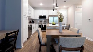 Una cocina moderna con armarios blancos, electrodomésticos de acero inoxidable y un comedor con mesa de madera y sillas tapizadas en gris.