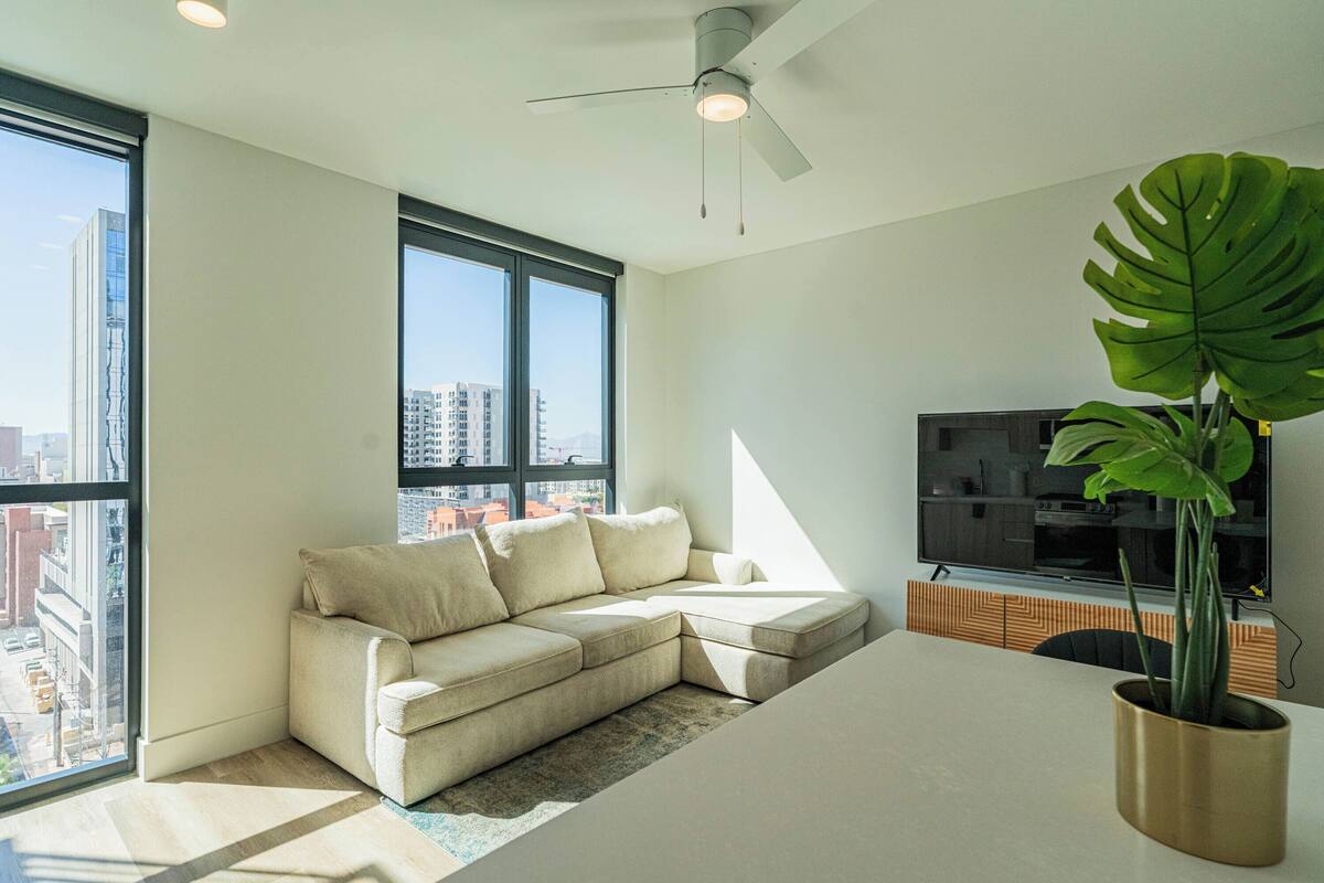 Een moderne woonkamer met een hoekbank, een flatscreen televisie, een plafondventilator en een groot raam met uitzicht op de stad.