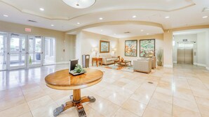 A spacious lobby with a round wooden table, a sofa, and a coffee table. There are doors leading to other areas, a painting on the wall, and an elevator. - Gorgeous, cozy beachfront condo with pool, pool spa & beach access (Perdido Key)