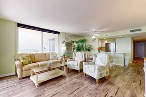 Ein geräumiges Wohnzimmer mit großem Fenster, einer Couch, zwei Sesseln, einem Couchtisch und einem Küchenbereich im Hintergrund.
