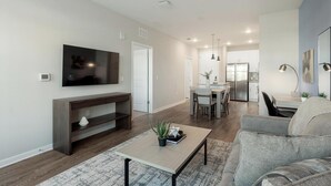 Una sala de estar moderna con un televisor de pantalla plana, un mueble de entretenimiento de madera, una mesa de centro con una planta, un sofá gris y un comedor con mesa y sillas.