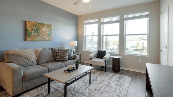 A living room with a gray sofa, a coffee table, and a chair. There is a painting on the wall and a window with blinds. - Landing | Spectacular 1BD, Gym, Yoga Studio (Four Corners)