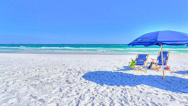 Una spiaggia con sabbia bianca, un cielo azzurro limpido e un ombrellone blu che offre ombra.