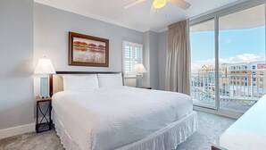 Ein Hotelzimmer mit einem großen Bett, zwei Nachttischen mit Lampen, einem Deckenventilator, einem großen Fenster mit Blick auf Gebäude und einem gerahmten Gemälde an der Wand.