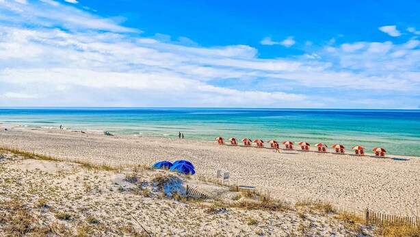 Uma praia com uma fileira de guarda-sóis vermelhos e brancos, areia dourada e o oceano ao fundo.