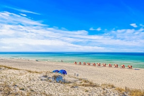 A beach with a row of red and white beach umbrellas, a sandy beach, and the ocean in the background. - Striking condo near the beach, pool, washer and dryer (Miramar Beach)