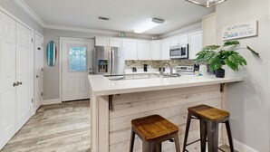 Uma cozinha com armários brancos, uma ilha grande, eletrodomésticos de aço inoxidável e uma placa com a inscrição 'Welcome to Beach House'.