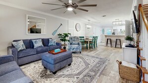 Uma sala de estar moderna com um sofá azul, uma mesa de centro e um tapete estampado. Há um ventilador de teto, um espaço de jantar com mesa e cadeiras, e uma cozinha com bancada e banquetas.