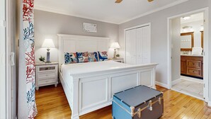 Quarto com uma cama grande, duas mesas de cabeceira com abajures, um ventilador de teto e uma mala no chão.
