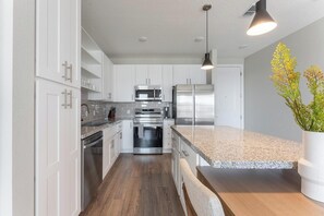 A modern kitchen with white cabinetry, stainless steel appliances, a granite countertop, and a hanging pendant light. - Landing | Stylish 2BD, Gym, Yoga Studio (Four Corners)