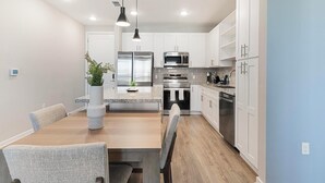 A modern kitchen with white cabinets, stainless steel appliances, and a wooden dining table with chairs. - Landing | Chic 2BD, Yoga Studio, Gym (Four Corners)