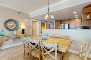 A dining area with a wooden table and chairs, a hanging light fixture, and a kitchen in the background. - Gulf-Front Condo On Okaloosa Island Playground, 2 Hot Tubs (Fort Walton Beach)