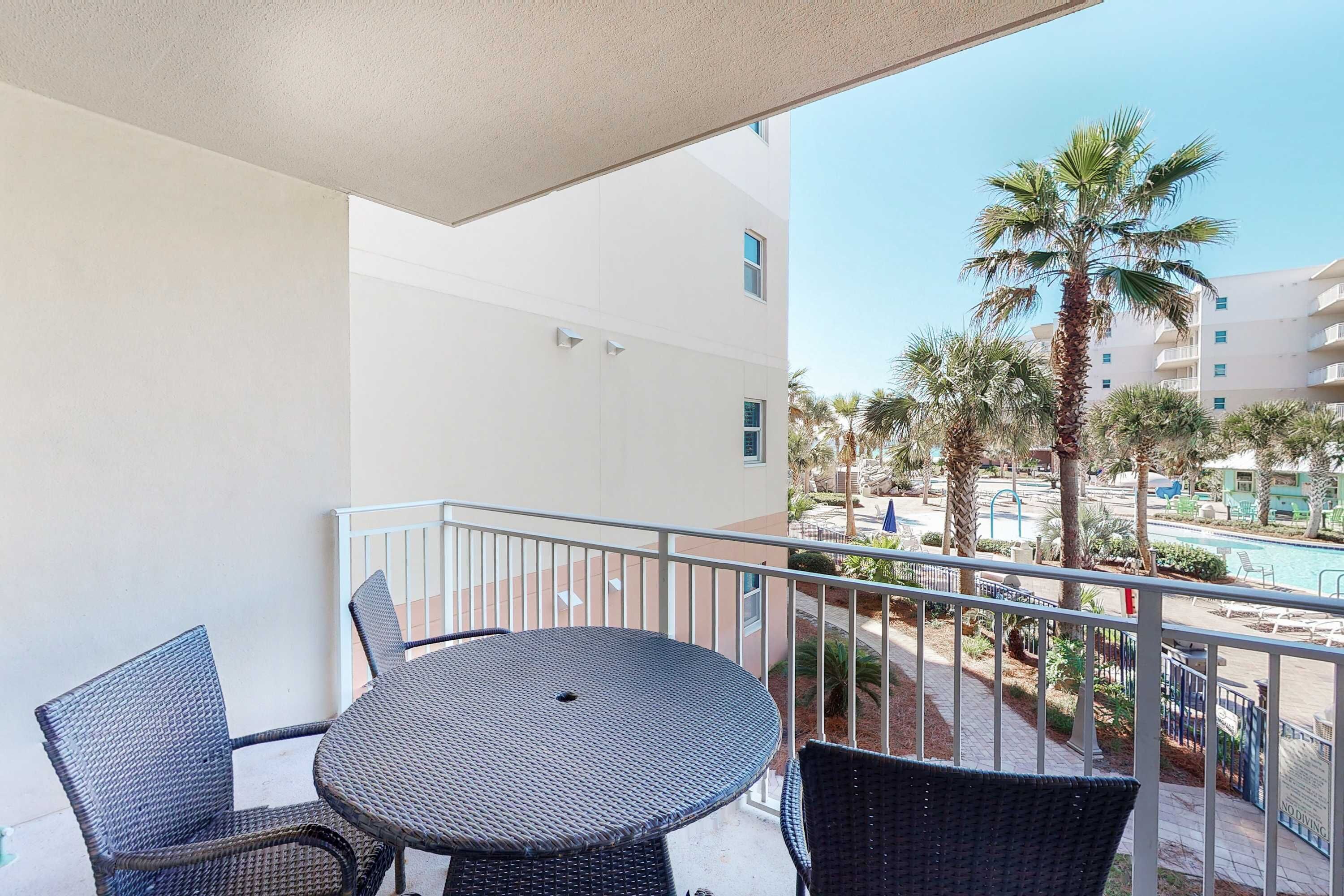 A balcony with a round table and four chairs, overlooking a pool area with palm trees and a building in the background.