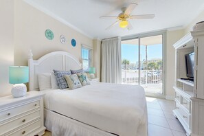 Ein Schlafzimmer mit einem großen Bett, einem Deckenventilator, einem Fernseher und einem Balkon mit Blick auf Palmen.