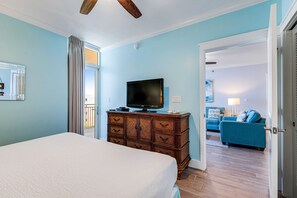 Ein Schlafzimmer mit einem Bett, einer Holzkommode mit Fernseher und Blick auf einen Wohnbereich mit einem blauen Sofa.
