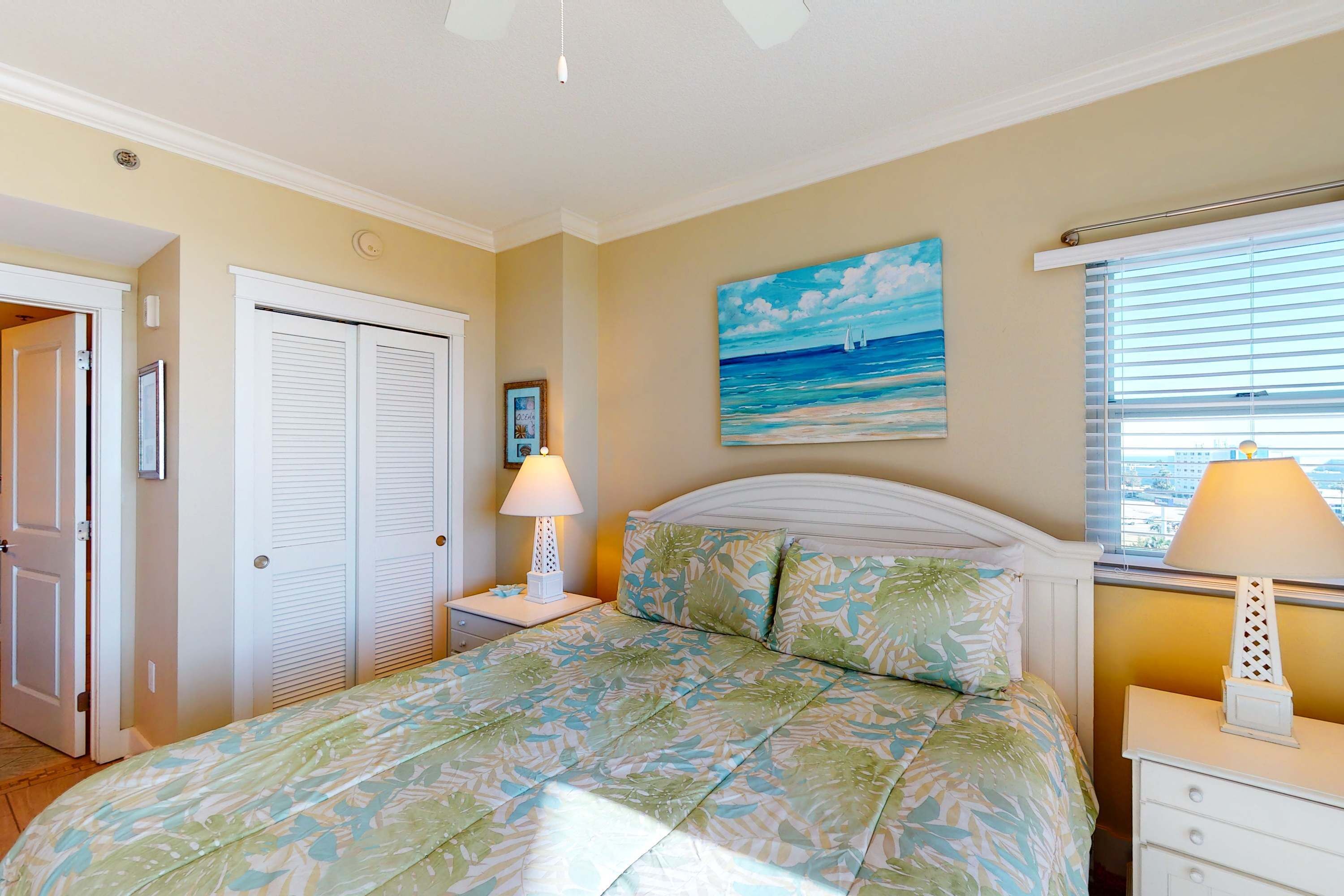 A bedroom with a bed, a nightstand, a window with blinds, and a painting of a beach scene.