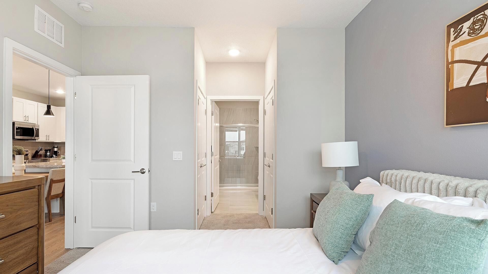A bedroom with a bed, bedside table, lamp, and a view of the bathroom and kitchen.