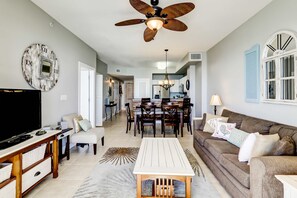 A spacious living room with a television, wooden coffee table, and a sofa. A dining area with a chandelier and chairs is visible in the background. - Gorgeous waterfront condo with incredible views, two pools & gym (Panama City Beach)
