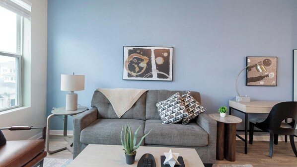 A modern living room with a grey sofa, a small side table with a lamp, a coffee table with a plant, and artwork on the wall. - Landing | Stunning 1BD, Yoga Studio, Gym (Four Corners)