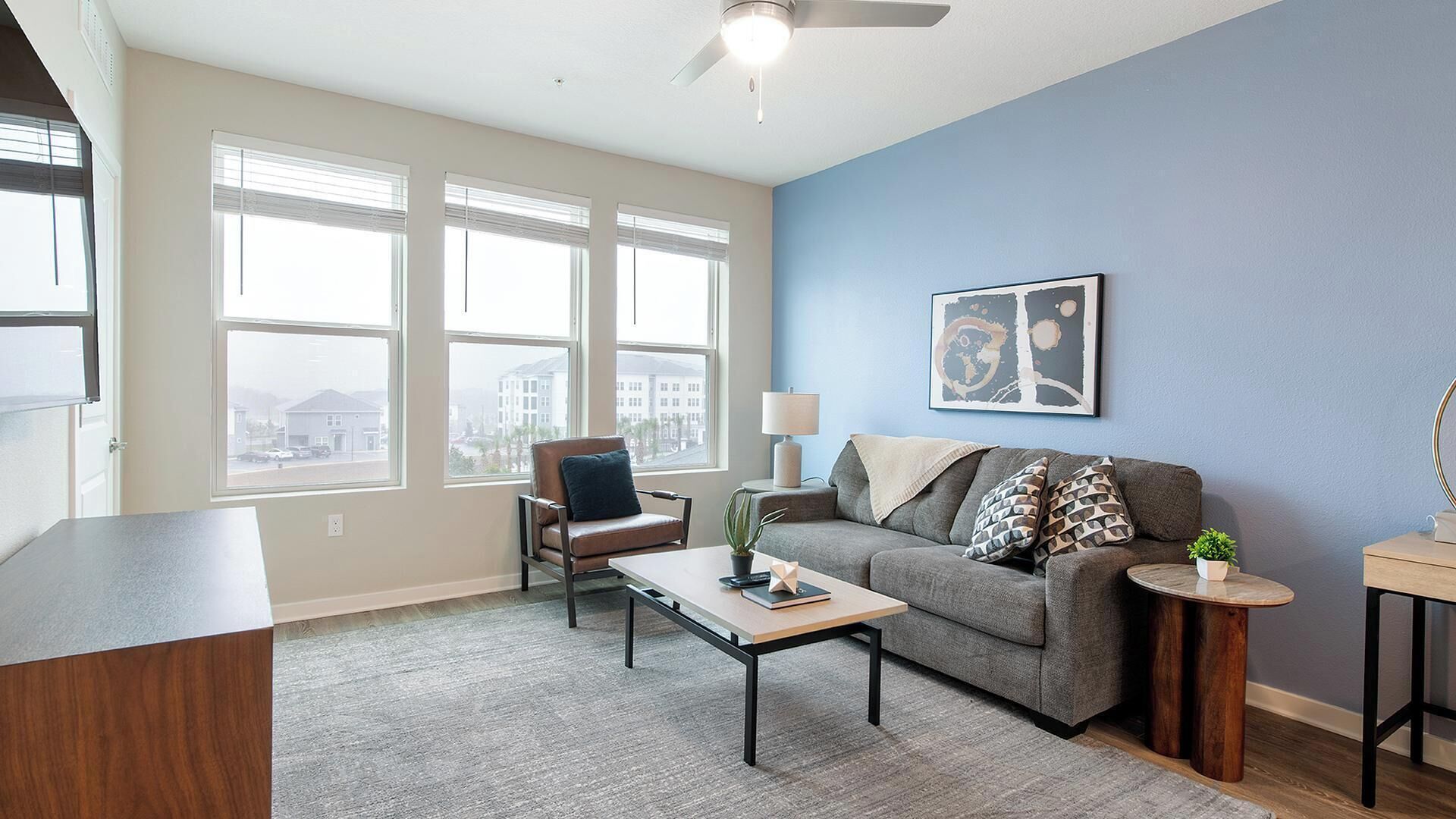 Ein modernes Wohnzimmer mit einem grauen Sofa, einem braunen Ledersessel, einem Couchtisch und einem Beistelltisch mit Pflanze. Große Fenster mit Jalousien, ein Deckenventilator und ein gerahmtes abstraktes Gemälde an der Wand.
