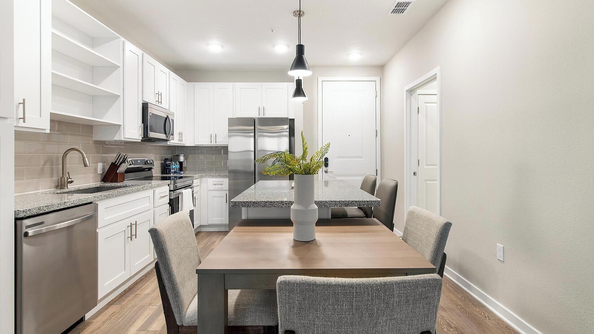 Una cocina moderna con armarios blancos, electrodomésticos de acero inoxidable y un comedor con mesa de madera y sillas grises.