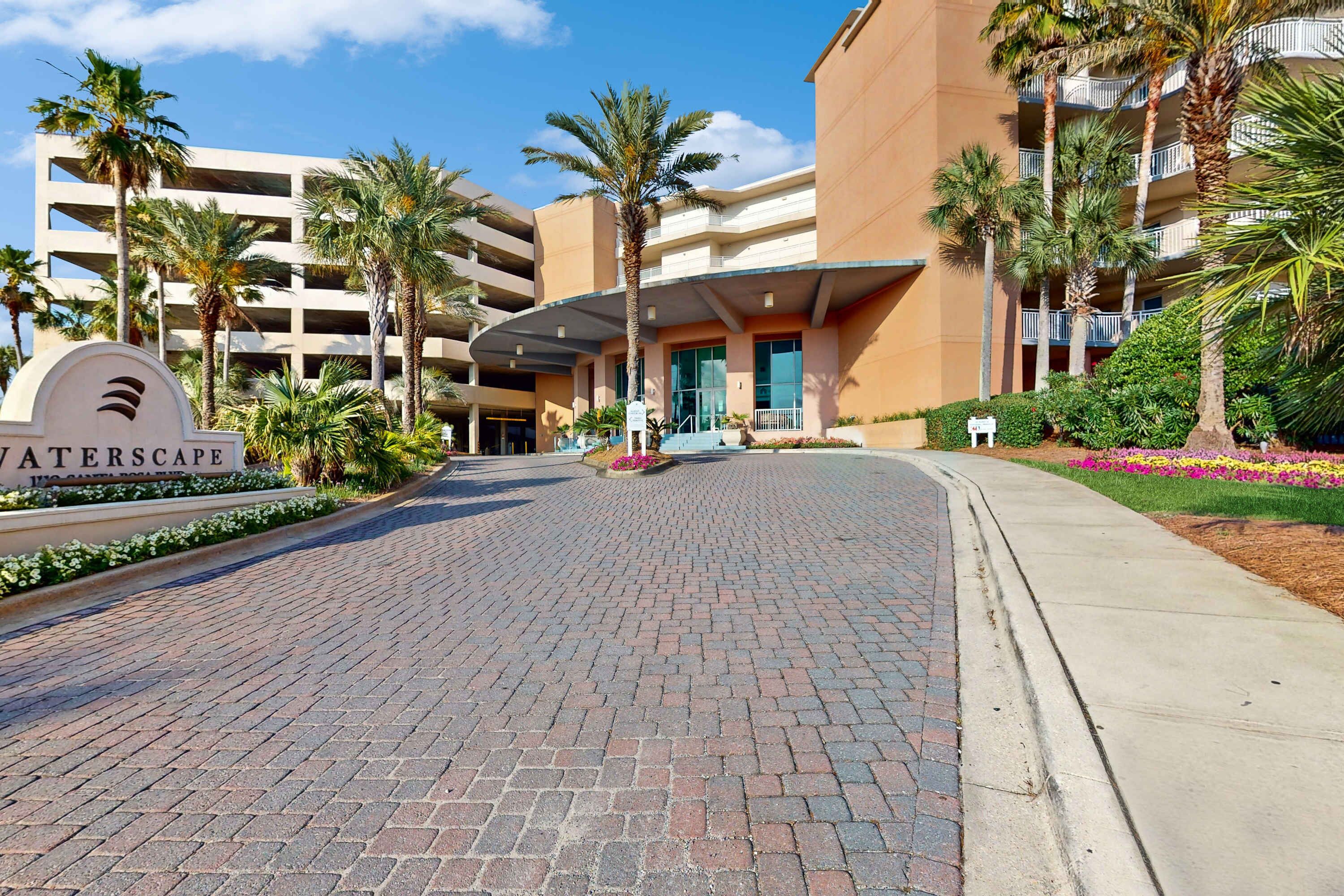 A hotel entrance with a sign reading "WATERSCAPE," featuring a brick pathway, palm trees, and a multi-story building with balconies.
