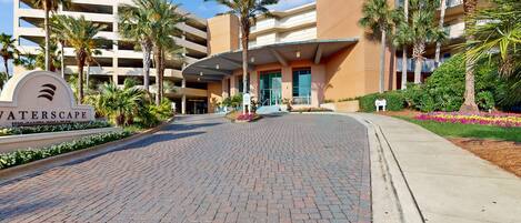 A hotel entrance with a sign reading "WATERSCAPE," featuring a brick pathway, palm trees, and a multi-story building with balconies.