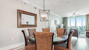 A dining area with a round table and six wicker chairs, a chandelier, and a large window with curtains. - Cozy, Tropical Condo On Okaloosa Island, Children's Playground, 2 Hot Tubs (Fort Walton Beach)