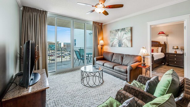 A hotel room with a ceiling fan, a sofa, a coffee table, a TV, and a large window with curtains. - Cozy, Tropical Condo On Okaloosa Island, Children's Playground, 2 Hot Tubs (Fort Walton Beach)