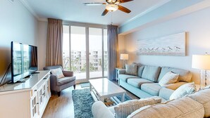 A modern living room with a flat-screen TV, a ceiling fan, a sofa, a coffee table, and a large window with curtains. - Elegant, Decorated Condo At Waterscape Waterfall, Lazy River (Fort Walton Beach)