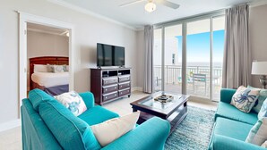 A hotel room with a balcony, a bed, a TV, and teal sofas. - Inviting, Spacious Condo On Okaloosa Island Waterfall, Lazy River (Fort Walton Beach)