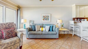 Un salon avec un canapé gris, un fauteuil à motifs, une petite table avec une lampe, et un coin cuisine avec des meubles blancs et un tabouret de bar.