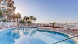 Une piscine entourée de chaises longues et de palmiers, le tout délimité par une clôture blanche et bordé d’un immeuble doté de balcons.