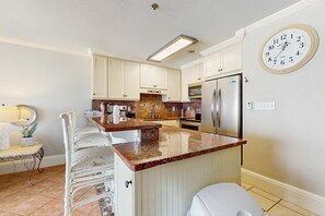 A kitchen with white cabinets, a granite countertop, a stainless steel refrigerator, and a wall clock. - Lovely beachfront condo with water views from private balcony + shared pool (Miramar Beach)