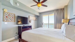 A hotel room with a large bed, a ceiling fan, a flat-screen TV, a wall-mounted artwork, and a view of the outside balcony. - Gulf-view condo with beach service, pools, hot tubs & gym (Panama City Beach)