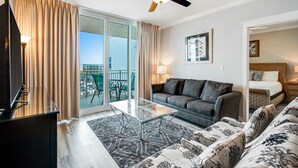 A hotel room with a sofa, a glass coffee table, a TV, a ceiling fan, and a balcony with a view. - 6th Floor Welcoming Condo At Waterscape 490 Feet Of Private Beach (Fort Walton Beach)