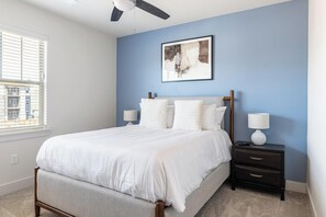 Une chambre à coucher comprenant un lit, deux tables de chevet, un ventilateur de plafond, une fenêtre avec des stores et un tableau abstrait accroché au mur.