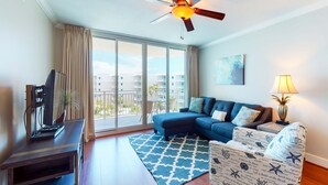 A modern living room with a ceiling fan, a sofa, a coffee table, a TV, and a large window with a view of buildings. - Warm, Welcoming Condo On Okaloosa Island 490 Feet Of Private Beach (Fort Walton Beach)