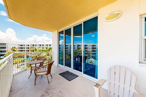 A balcony with a table and chairs, a view of a swimming pool, and a building with balconies in the background. - Warm, Welcoming Condo On Okaloosa Island 490 Feet Of Private Beach (Fort Walton Beach)