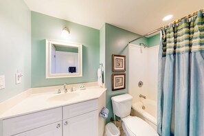 A bathroom with a white sink, mirror, and toilet, a shower curtain, and framed pictures on the wall. - Warm, Welcoming Condo On Okaloosa Island 490 Feet Of Private Beach (Fort Walton Beach)