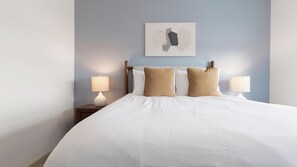 Un letto rifatto con lenzuola bianche e due cuscini color beige, affiancato da due comodini con lampade, su una parete azzurra con un'opera d'arte geometrica sopra.