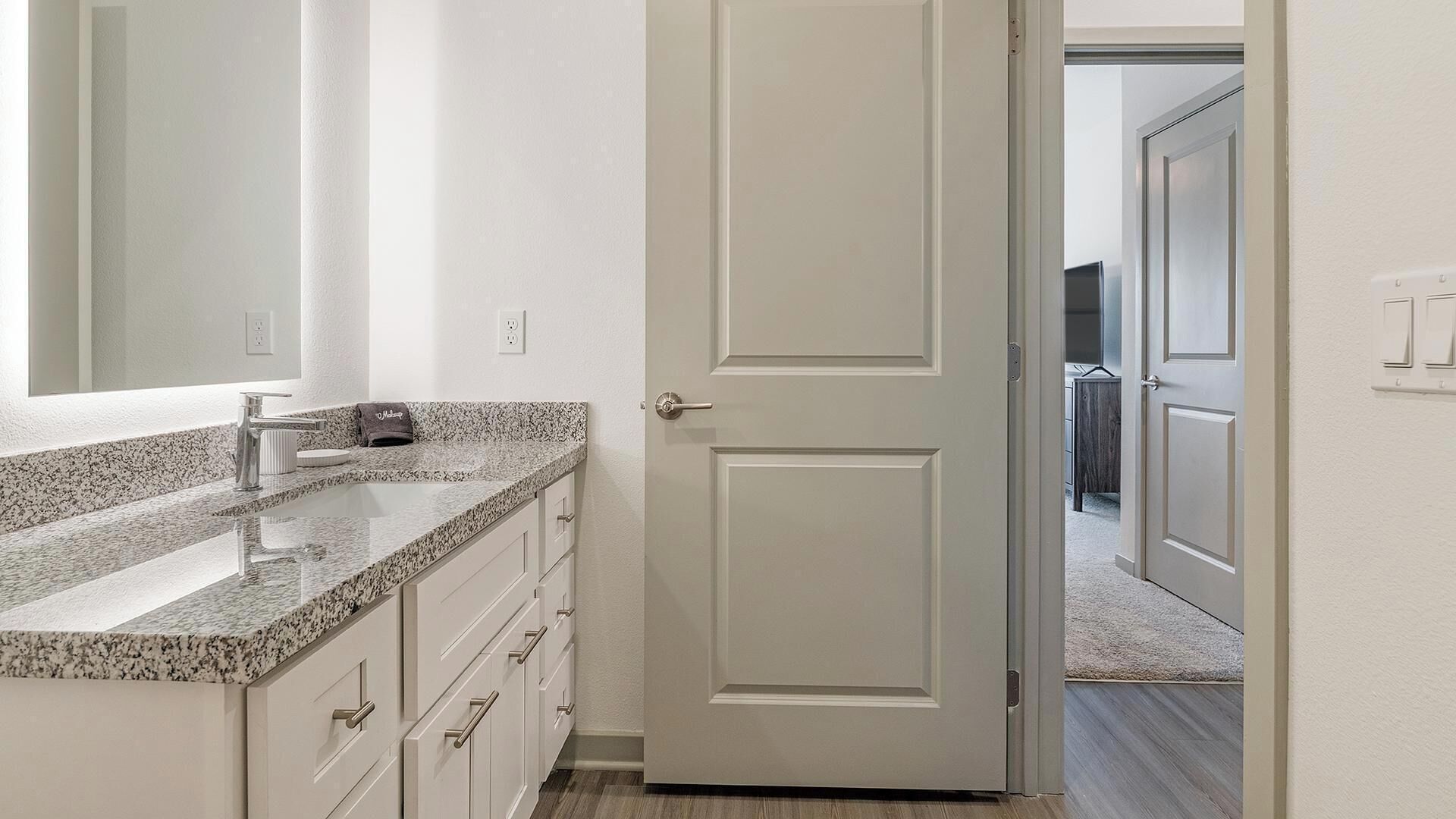 A bathroom with a granite countertop, a mirror, a towel, and a door leading to another room.