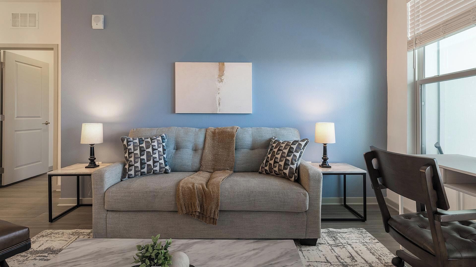 Ein Wohnzimmer mit grauem Sofa, zwei Nachttischen mit Lampen, einem Couchtisch, einem Sessel und einem Fenster mit Jalousien.