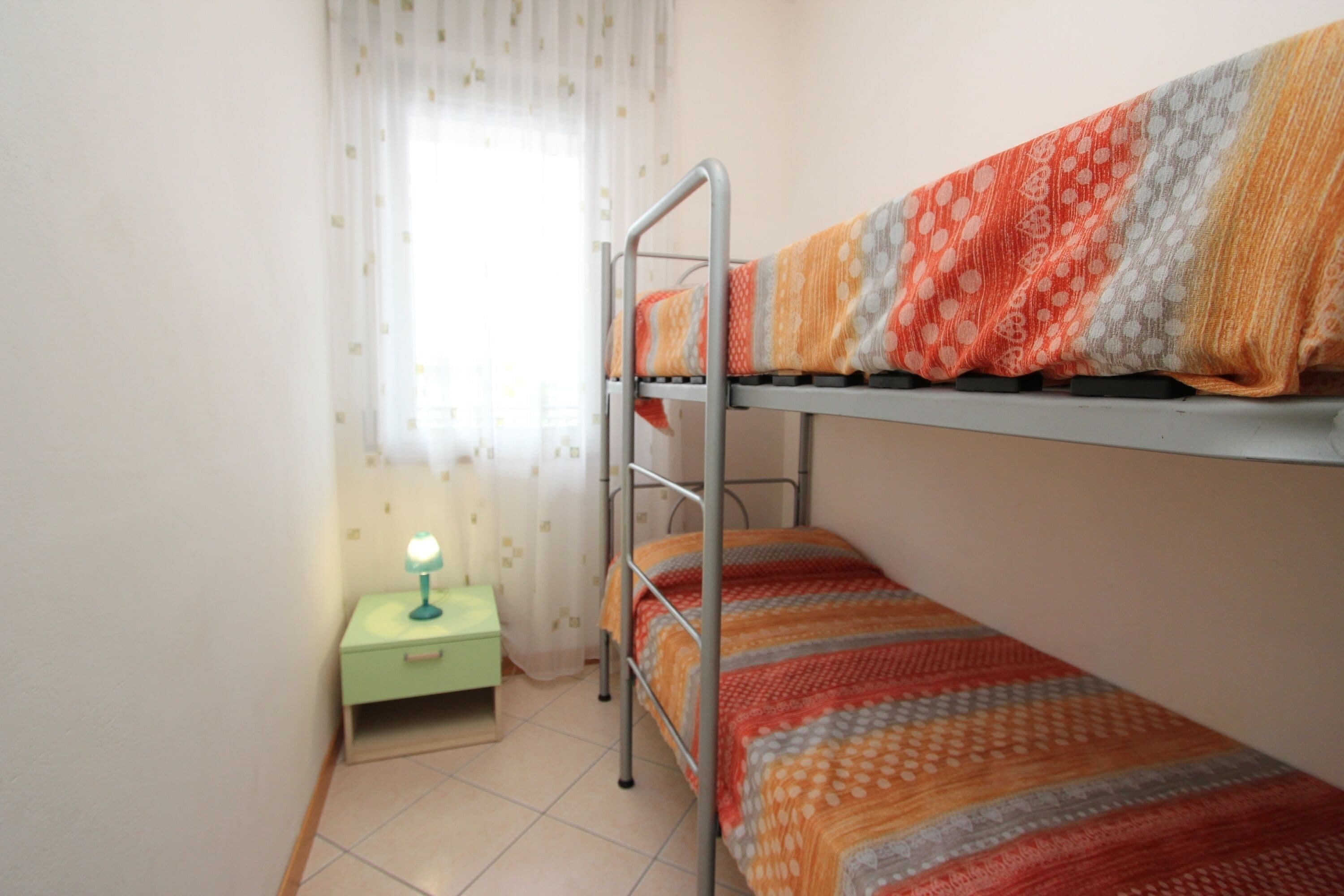 A bunk bed with a patterned blanket, a small bedside table with a lamp, and a window with curtains.