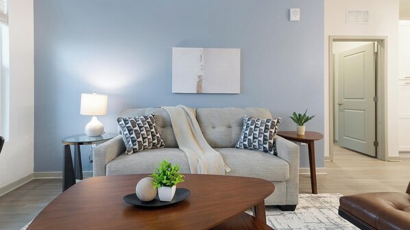 Ein Wohnzimmer mit grauem Sofa, gemusterten Kissen, einem Holztisch, einem kleinen Beistelltisch mit Lampe und einem Blumentopf.