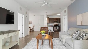 Una sala moderna con televisor de pantalla plana, un comedor con mesa y sillas de madera, una cocina con armarios de madera oscura y electrodomésticos de acero inoxidable, y un ventilador de techo.