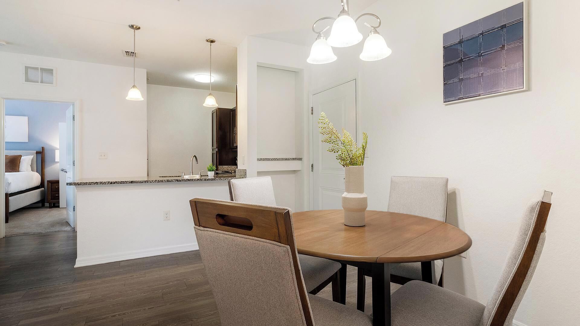 A dining area with a round table, chairs, pendant lights, a vase with flowers, and a painting on the wall.