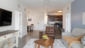 A modern living room with a flat-screen TV, a dining area with a table and chairs, a kitchen with appliances, and a gray sofa. - Landing | Chic 2BD, Game Room, Clubhouse (Apopka)
