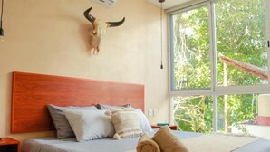 A bedroom with a wooden headboard, a large window with a view of greenery, and a bed with a beige blanket and pillows. - Gama 09 Apartments - Apartment with balcony (Tulum)